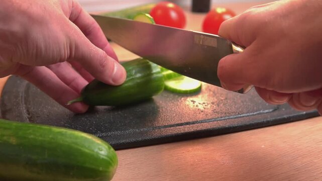  knife cutting crisp green cucumber into thin rounds on black cutting board nearby whole cucumbers and tomatoes healthy vegetable slicing salad prep fresh organic detail