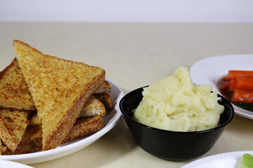 Mashed potatoes in a black bowl and several slices of cheese bread on a plate on a light-colored dining table.