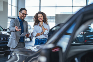 Professional Car Dealership Consultation With Smiling Couple Reviewing Tablet In Showroom During...