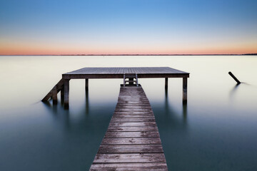 Silent Deck. Longue exposition réalisée au lac de Biscarrosse, où un ponton en bois avance lentement vers une plateforme posée sur l’eau calme. 
