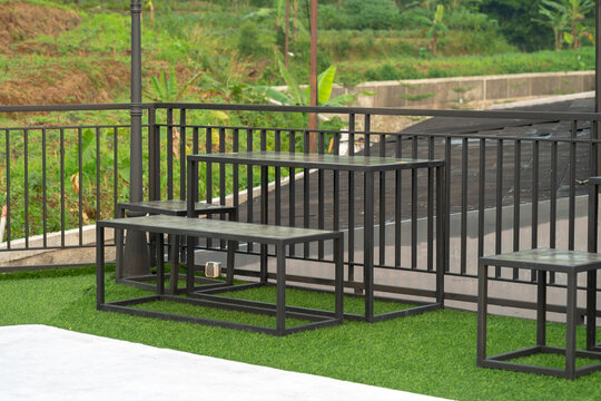 Minimalist black metal tables and benches on artificial grass at an outdoor terrace. A modern industrial seating area overlooking a green hillside, perfect for a relaxing coffee break - Powered by Adobe