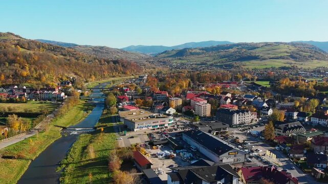 Aerial view of Mszana Dolna town center nestled in the Beskidy Mountains, Poland. Scenic autumn panorama featuring residential areas, river, and forested rolling hills.