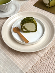 A slice of matcha roll cake served on a white ceramic plate with green tea and wooden cutlery on a beige cloth.Bright,clean tabletop food photography ideal for dessert menus and bakery promotions.