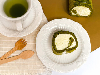 A slice of matcha roll cake served on a white ceramic plate with green tea and wooden cutlery on a beige cloth.Bright,clean tabletop food photography ideal for dessert menus and bakery promotions.