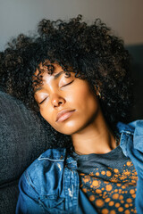 A serene woman with curly hair relaxes with her eyes closed, embodying calmness and tranquility against a cozy background.