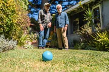 Two men stand on a grassy lawn, observing a blue ball in the foreground, surrounded by greenery and a house in the background.
