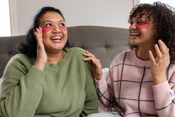 Female friends laughing together on bed in bedroom while wearing red gel under-eye patches