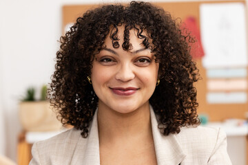 Woman wearing blazer smiling at camera in office with desk, succulent plant and cork bulletin board