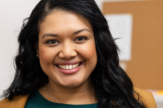 Business woman wearing brown blazer smiling while standing in office with corkboard and necklace