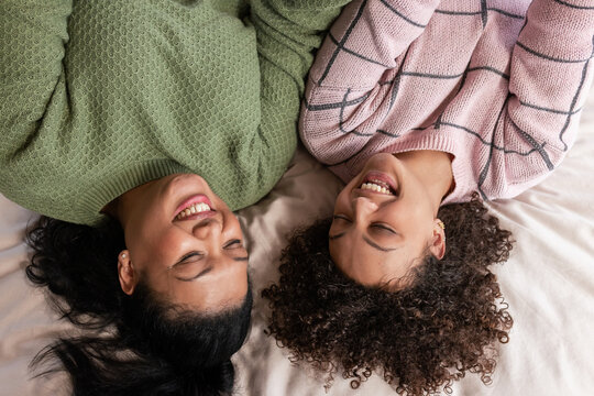 Female friends lying on cream bed sheets in bedroom smiling wearing green and pink knit sweaters