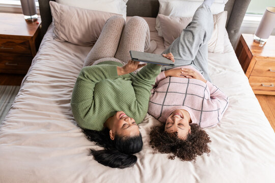 Two women lying side by side near headboard sharing tablet on bedding with nightstands in bedroom