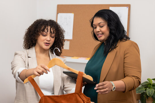 Female fashion designers examining rust-colored garment on hanger in design studio with tablet - Powered by Adobe