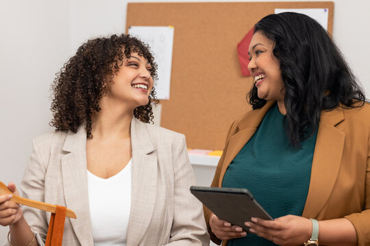 Two female colleagues exchanging ideas at office by cork board with fabric sample and tablet