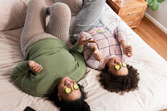 African American mother and daughter relaxing on bed in bedroom with cucumber slices and smartphone