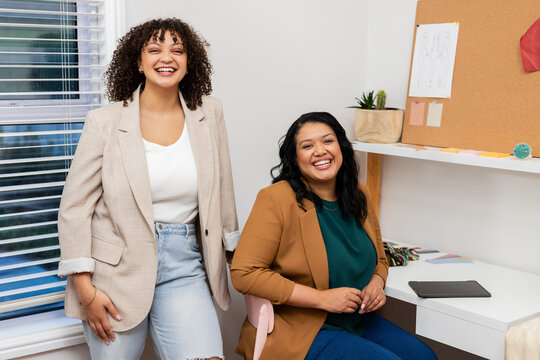 Female coworkers wearing blazers discussing design ideas at office desk with tablet and cork board