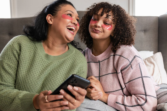 African American mother and daughter sharing tablet, wearing under-eye gel patches, lounging on bed - Powered by Adobe