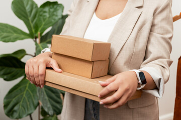 Woman in blazer holding three stacked cardboard shipping boxes in home office checking smartwatch
