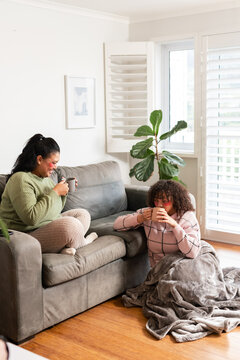 Diverse mother and daughter chatting while holding mugs on sofa at home beside fiddle leaf fig