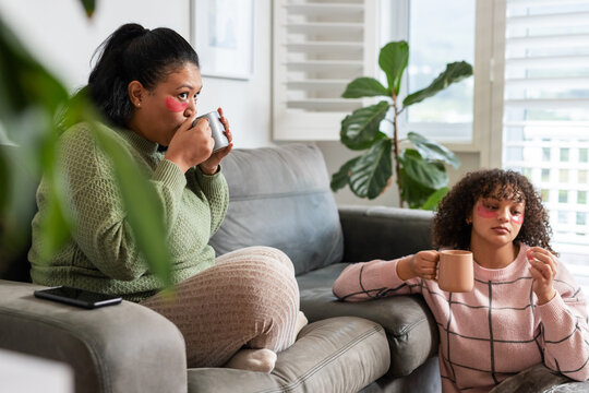 African American mother and daughter wearing gel patches sipping from mugs on sofa with phone