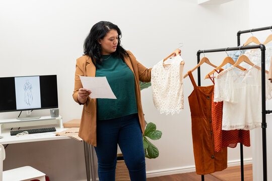 Woman examining lace top on hanger while holding printed design sheet in fashion design studio