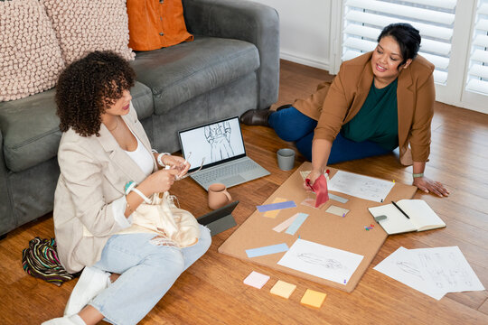 Female coworkers collaborating with fabric swatches on floor in living room with cork board, laptop
