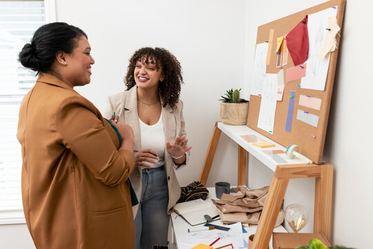 African American women coworkers standing at table reviewing fabric swatches on corkboard in studio