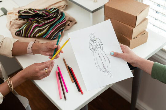 Diverse female coworkers reviewing fashion sketch sheet at desk by window selecting colored pencils