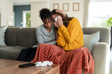 Diverse female friends comforting each other on sofa in living room with crumpled tissues