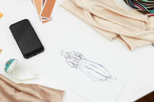 Fashion sketch resting on white desk with phone, color cards, pin cushion, beige striped fabrics