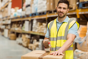 Man wearing hi-vis vest sealing cardboard box in depot amid racks with tape dispenser, copy space © wavebreak3