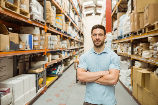Man in late 20s wearing light blue polo standing in warehouse aisle among shelving and boxes - Powered by Adobe