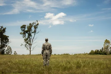 Fotobehang Beren Male soldier standing with back to horizon in grassy field wearing camouflage uniform and cap  © wavebreak3