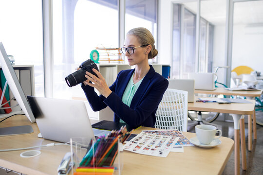 Female creative wearing navy blazer examining DSLR camera, sitting in office with laptop, prints