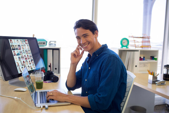 Chinese adult male sitting at office desk wearing blue shirt editing headshots on laptop with DSLR - Powered by Adobe