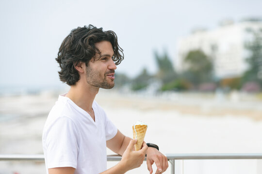 Man leaning on beach railing holding waffle cone wearing white V-neck tee and wristwatch