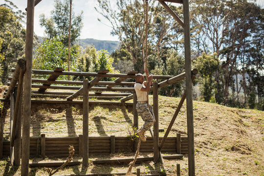 African American man climbing rope on obstacle on grassy slope in camo pants, copy space - Powered by Adobe