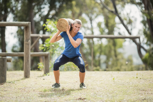 Woman holding wooden log on right-shoulder squatting in grassy area with posts, rails in blue shirt