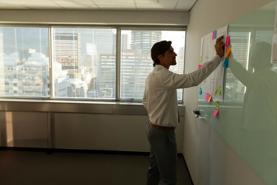 Mid-adult man organizing sticky notes and printed sheets on glass whiteboard in office, copy space
