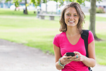 Woman smiling holding smartphone on park path wearing tee backpack strap bracelets ring, copy space