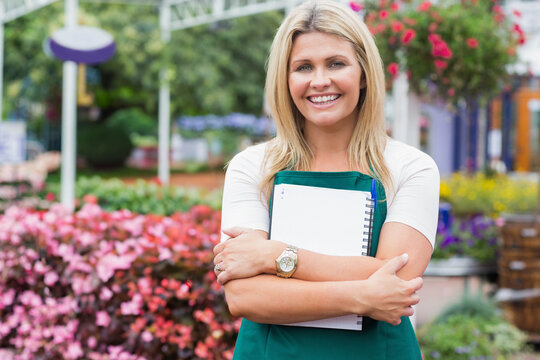 Woman wearing green apron standing in plant nursery holding spiral-bound notebook and smiling