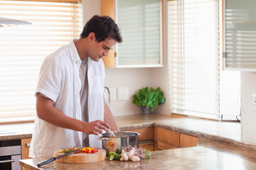Man in his 20s stirring pot while prepping peppers on cutting board at kitchen island