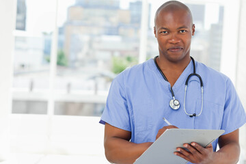 Adult African-American man writing on clipboard with pen in clinic in blue scrubs and stethoscope
