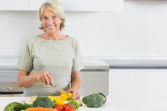 Senior woman slicing yellow bell pepper on wood board with chef knife in bright kitchen