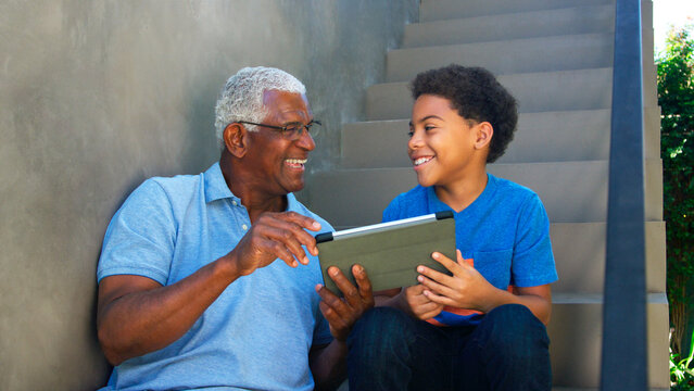 Grandfather With Grandson Sitting On Steps Outdoors At Home Using Digital Tablet