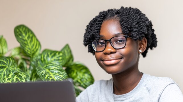 A smiling girl with curly hair and glasses sits at a laptop, surrounded by greenery, exuding a confident and cheerful demeanor.