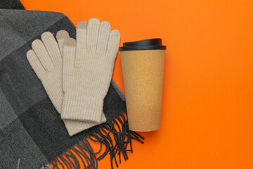 Cozy Autumn Scene: Warm Knitted Gloves, Plaid Scarf, and Reusable Coffee Cup on Bright Orange Background