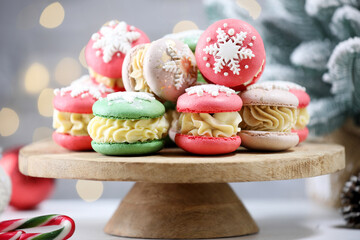 Tasty Christmas macarons on wooden stand against blurred lights, closeup