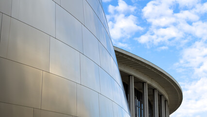 Fototapeta premium LOS ANGELES, CA, OCT 10, 2025: Smooth curved metallic wall of Walt Disney Concert Hall meets traditional concrete structure in background, with columned overhang under bright cloudy sky