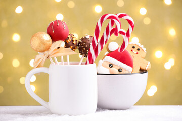 Tasty cake pops, candy canes and gingerbread cookies on white table against beige background with blurred lights. Christmas treat