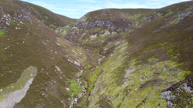 Aerial view of a stream winding through a valley, its banks adorned with green and purple vegetation, contrasting with the rocky terrain, Brechin, Scotland, United Kingdom.